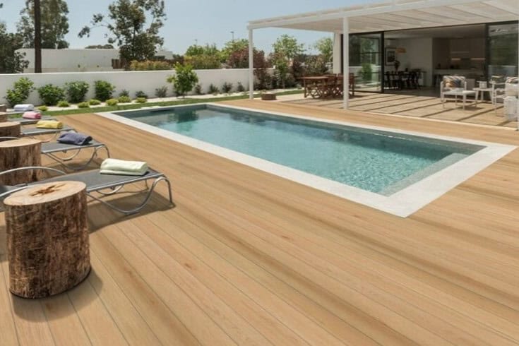 The wooden deck, sourced from a renowned deck materials supplier, features a rectangular swimming pool surrounded by lounge chairs and tree stump tables. A covered patio with seating is visible in the background, bordered by lush plants and trees.