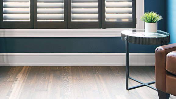 Room corner with wooden floor, blue walls, a round glass side table with a plant, and brown shutters on the window.