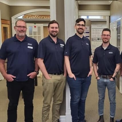 Four men stand side by side in a hallway, wearing matching navy blue shirts with name tags. They are smiling and posing for the photo.