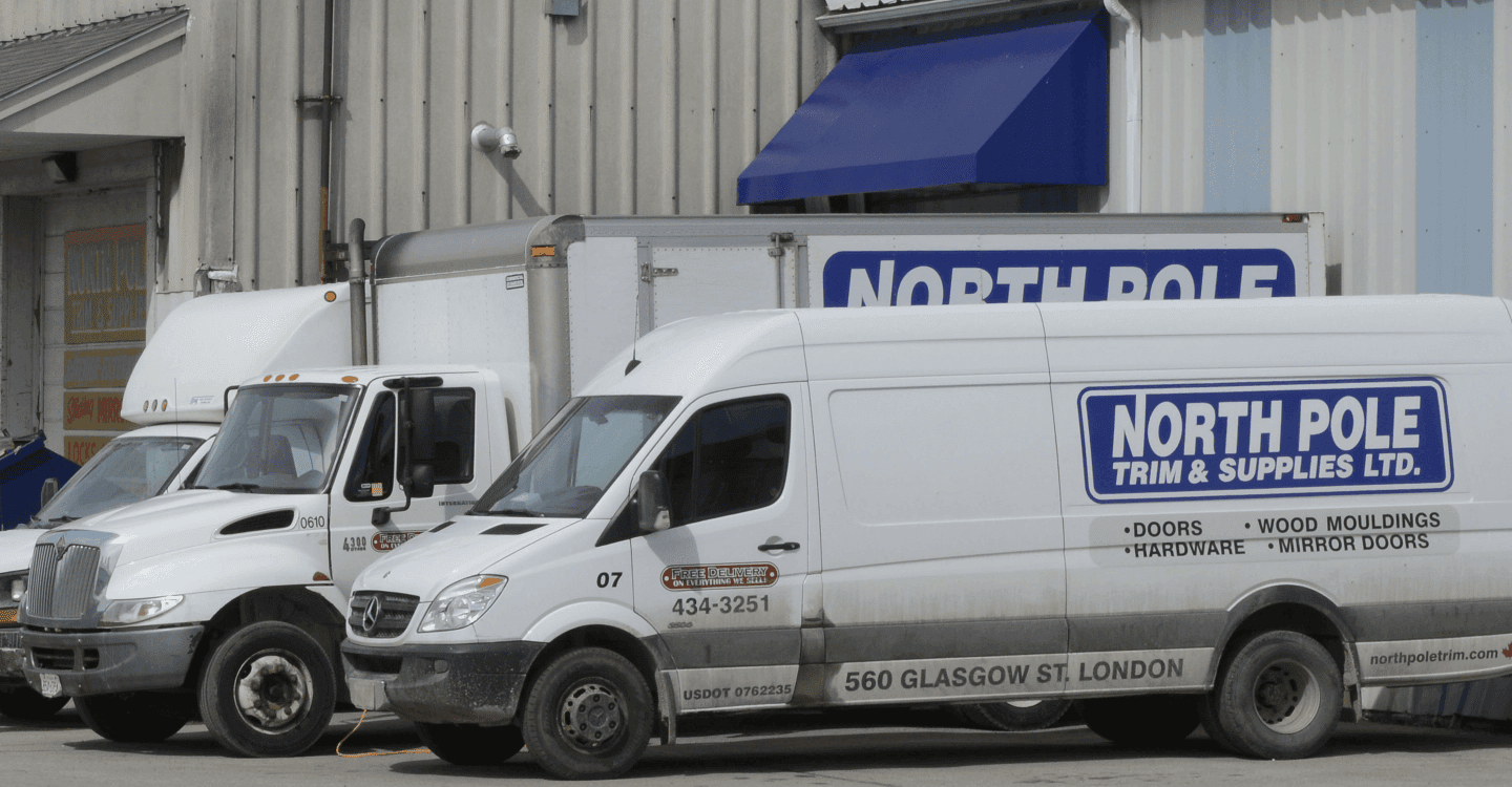 Two delivery trucks parked outside a warehouse under a blue awning, both branded with "North Pole Trim & Supplies Ltd." A sign with the same branding is visible above the door.