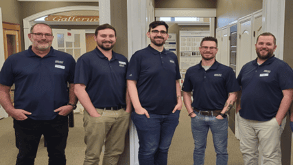 Five men wearing matching navy blue polo shirts with name tags stand in a hallway, smiling at the camera, ready to offer you a free quote.