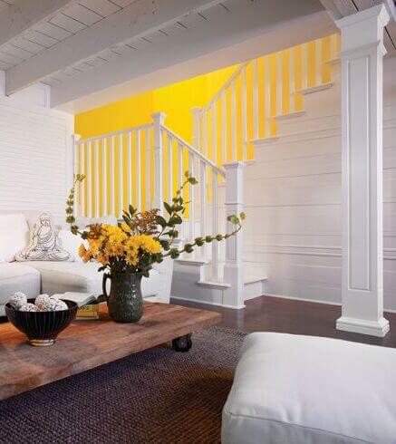 A cozy living room with a wooden coffee table, a flower vase centerpiece, a white sofa, and a staircase with a bright yellow accent wall.