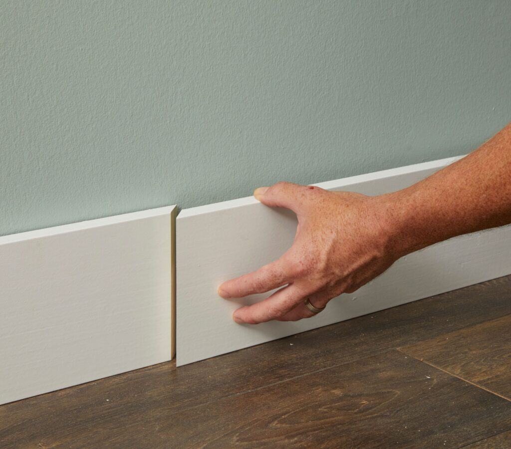 A person adjusts a white baseboard on a wall with light green paint above a dark wood floor.