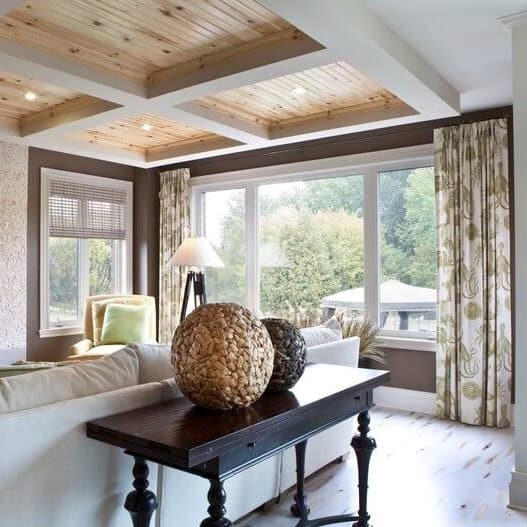 A living room features a white couch, a dark wood table with decorative woven spheres, patterned curtains, large windows, and a wooden coffered ceiling.