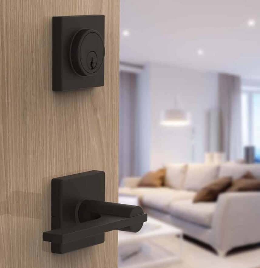 Close-up of a modern black door handle and lock on a wooden door, slightly ajar, revealing a blurred view of a living room with a sofa and lamp in the background.