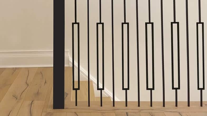 Black metal railing with vertical bars and rectangular accents on a light wood floor and white wall background.