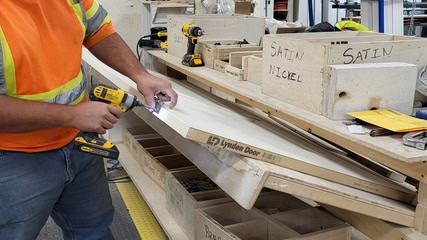 A person in an orange safety vest uses a power drill on a pre-hung door component. Nearby, boxes labeled Satin Nickel contain tools and materials.