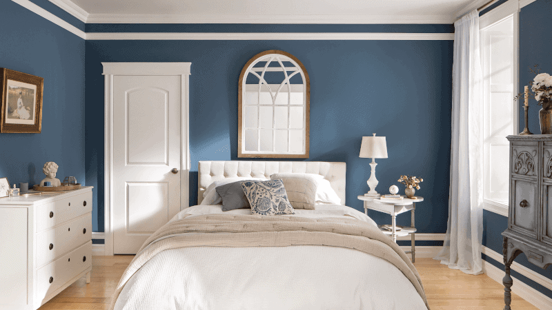 Bedroom with blue walls, a white bed, a round mirror above the bed, a dresser, a nightstand with a lamp, and a window with sheer curtains.