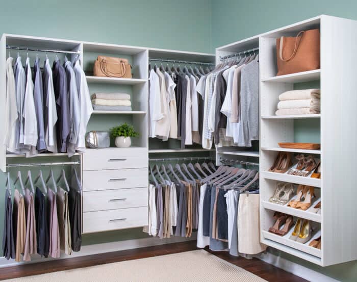 A neatly organized walk-in closet with assorted clothing on hangers, shelves with shoes, bags, towels, and drawers. Walls are painted green and flooring is dark wood.