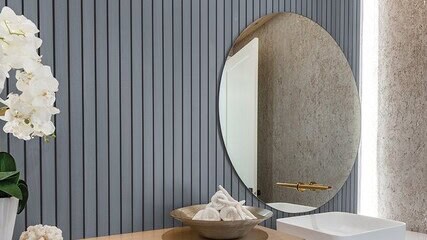 Bathroom scene with a round mirror, a white sink with a gold faucet, a bowl with white towels, and a potted orchid on a wooden countertop. Vertical gray panel wall in the background.