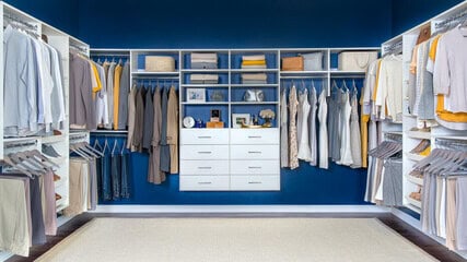 Spacious walk-in closet with neatly organized clothes in neutral tones, shelves with various items, and white cabinets against a blue wall.